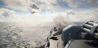 Naval ship firing a cannon with a fighter jet flying overhead