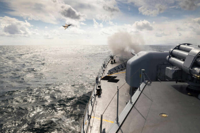 Naval ship firing a cannon with a fighter jet flying overhead
