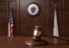 A wooden gavel resting on a table in a courtroom with flags in the background