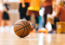 A basketball resting on the gym floor with players blurred in the background
