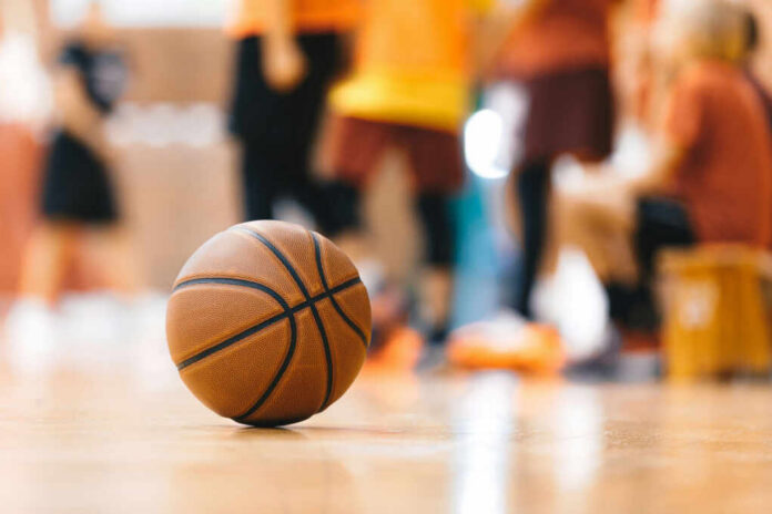 A basketball resting on the gym floor with players blurred in the background