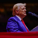 A man in a blue suit and red tie speaking into a microphone on stage