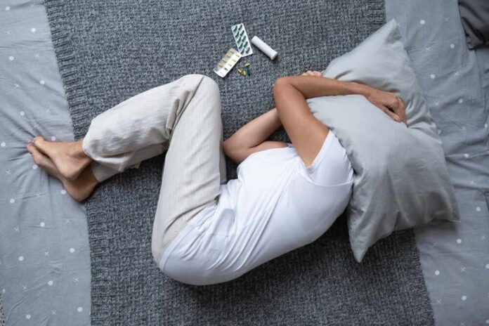 Person lying on a bed with a pillow over their head, surrounded by medication