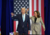 President Biden and Vice President Harris waving at a political event with an American flag backdrop