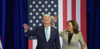President Biden and Vice President Harris waving at a political event with an American flag backdrop