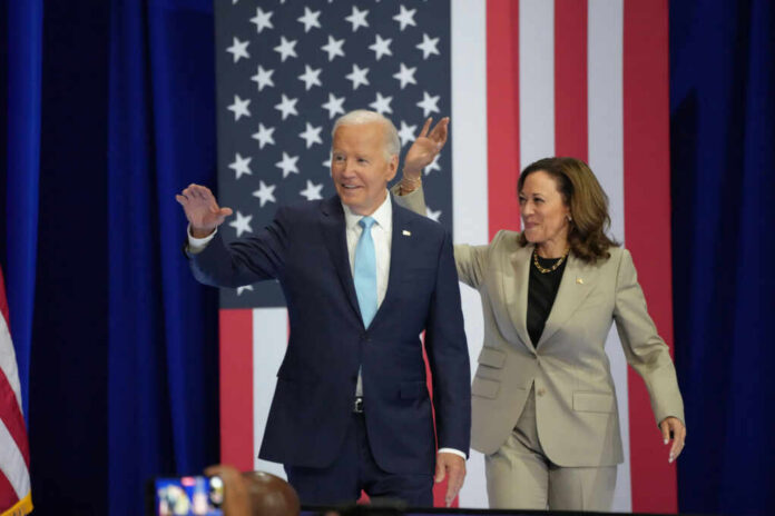 President Biden and Vice President Harris waving at a political event with an American flag backdrop