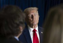 A serious-looking political figure in a suit and red tie during a media interaction