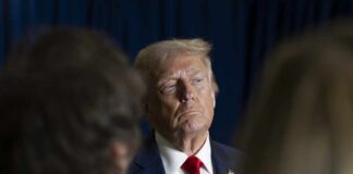 A serious-looking political figure in a suit and red tie during a media interaction