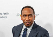 A man in formal attire smiling at a media event with a branded background