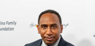 A man in formal attire smiling at a media event with a branded background