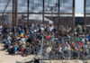 Large group of migrants sitting near a border fence with law enforcement present