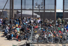 Large group of migrants sitting near a border fence with law enforcement present