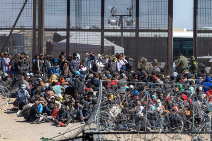 Large group of migrants sitting near a border fence with law enforcement present