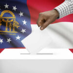 TRUMP Endorsement Swings Georgia Runoff A hand placing a ballot into a ballot box with the Georgia state flag in the background