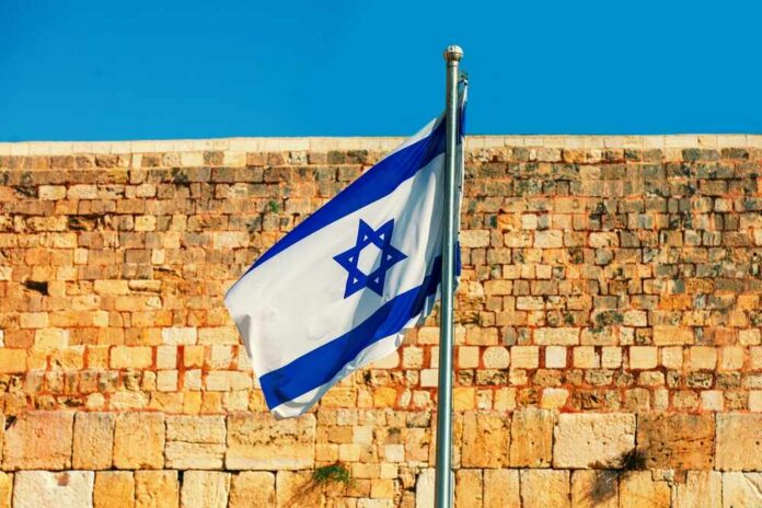 Israeli flag waving in front of a stone wall under a clear blue sky