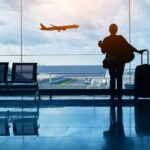Silhouette of a traveler holding a coffee cup and standing next to a suitcase, looking out at an airplane taking off from the airport