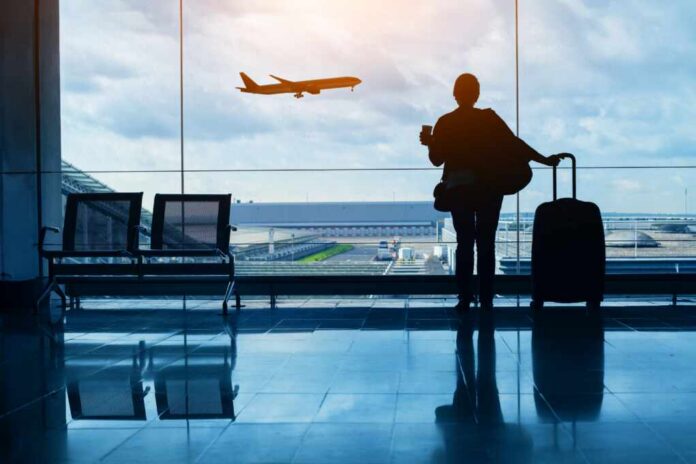 shutterstock_2467550689.jpg Silhouette of a traveler holding a coffee cup and standing next to a suitcase, looking out at an airplane taking off from the airport