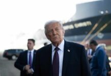 A political figure in a suit looking thoughtfully at an outdoor event with a ship in the background