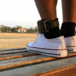 Fake Heiress Turns Courtly Chain Into Fashion Trend A persons foot wearing a white sneaker with an ankle monitor, sitting on a park bench