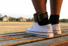 A persons foot wearing a white sneaker with an ankle monitor, sitting on a park bench