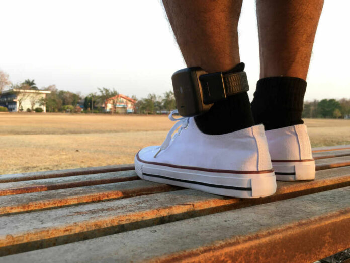 A persons foot wearing a white sneaker with an ankle monitor, sitting on a park bench