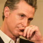 DHS SLAMS Newsom: “Releasing Murderers!” Close-up of a man in a suit with a thoughtful expression, hand on chin