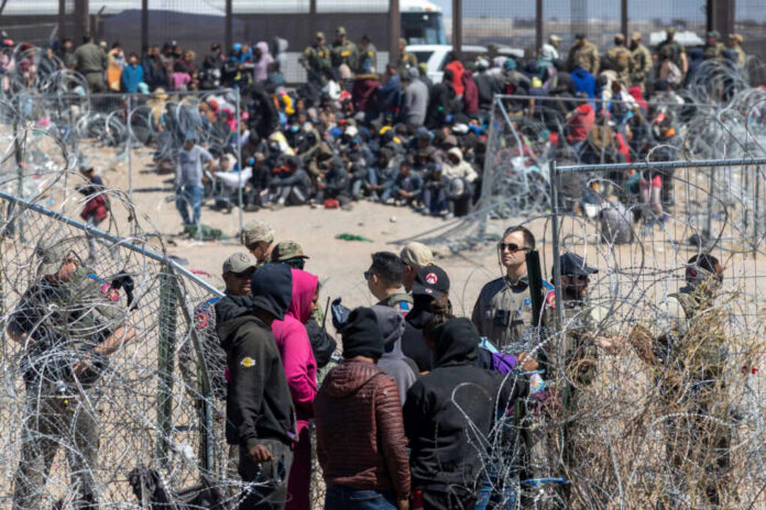 A crowd of people at a border area with law enforcement and barbed wire