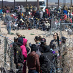 A crowd of people at a border area with law enforcement and barbed wire
