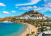 A picturesque coastal view featuring a beach, a historic castle on a hill, and a Mediterranean village