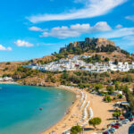 A picturesque coastal view featuring a beach, a historic castle on a hill, and a Mediterranean village