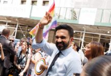 Smiling man waving at a pride parade with a rainbow flag in the background