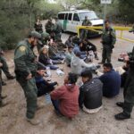 Border agents surround a seated group near a van