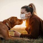 Woman in mask touching noses with brown dog at sunset