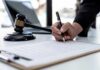 Hand signing legal document beside gavel and laptop