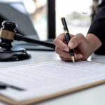 Hand signing legal document beside gavel and laptop