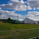 Temporary white tents and equipment on a grassy field with traffic cones and metal barriers
