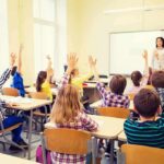 Students raise hands in a classroom with a teacher at the front