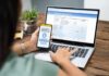 Person using smartphone to approve bank transfer beside laptop with online banking
