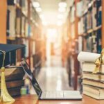 Graduation cap on books beside laptop and diploma in library aisle