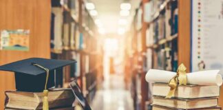 Graduation cap on books beside laptop and diploma in library aisle