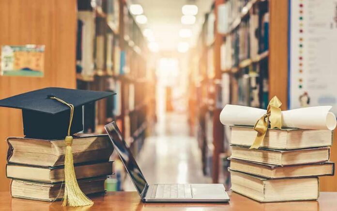 Graduation cap on books beside laptop and diploma in library aisle